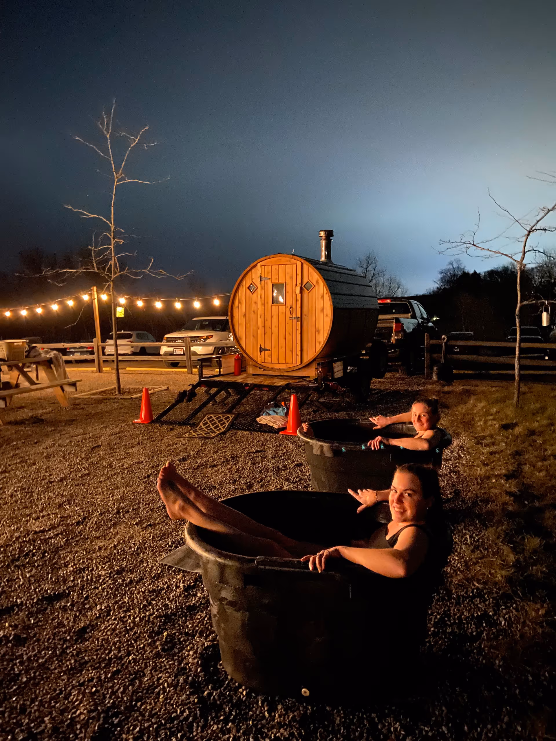 Night scene shows outdoor tubs with people relaxing near a wooden barrel sauna and string lights.