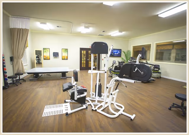 Well-lit interior fitness/therapy room with exercise machines, a treatment table, and wood floors.