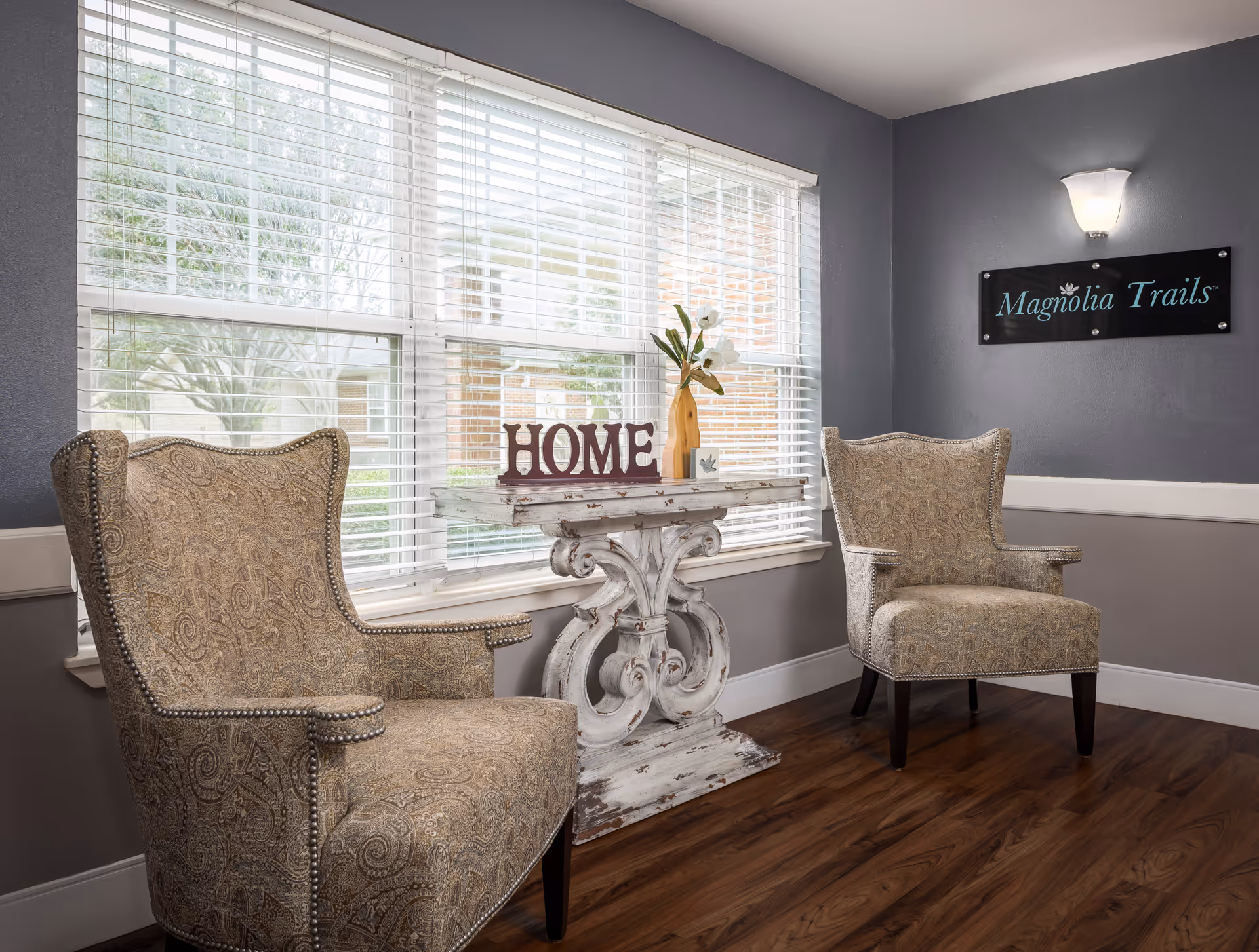 A cozy seating area with two patterned armchairs facing each other, separated by a distressed white wooden table with decorative scrollwork. On the table is a vase with white flowers and a decorative sign spelling 'HOME'. Behind the table is a large window with white blinds, and on the gray wall to the right is a sign that reads 'Magnolia Trails'. The floor is a warm wood tone.