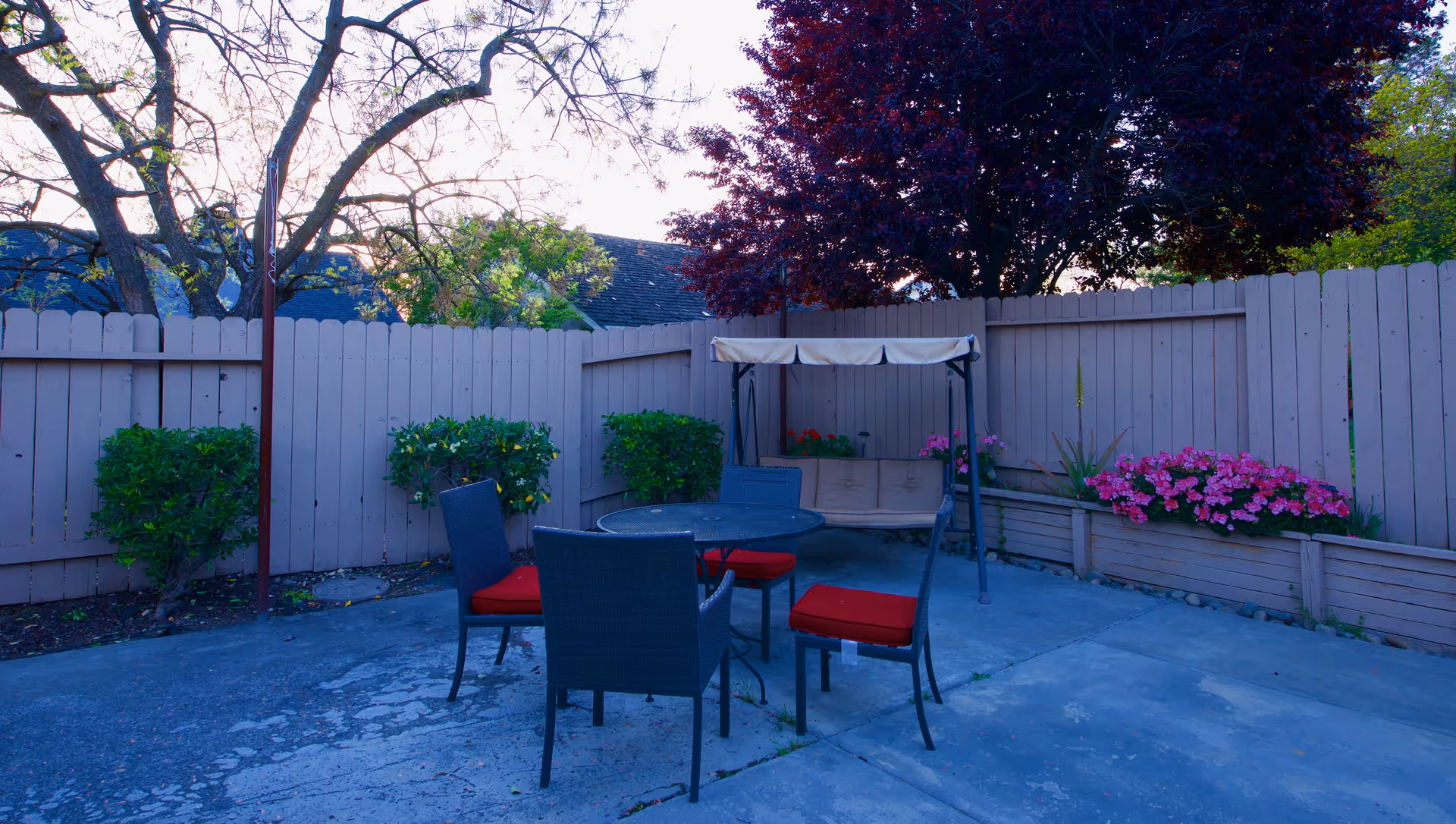 Backyard patio with table, chairs, a swing bench, and flowering plants along a fence.