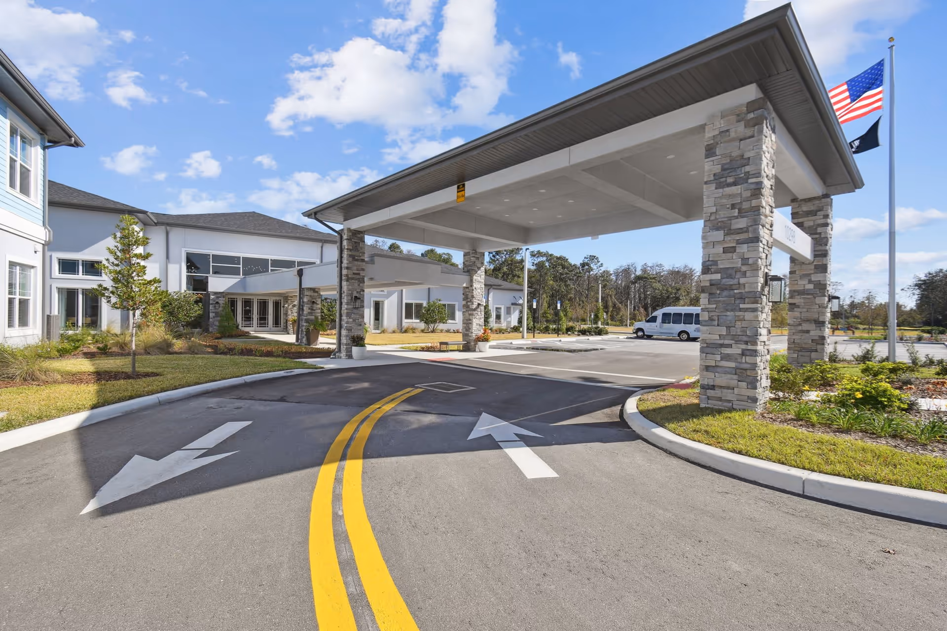 Covered entrance/porte-cochère and driveway of a senior living facility with stone columns, flags, and a parked van.