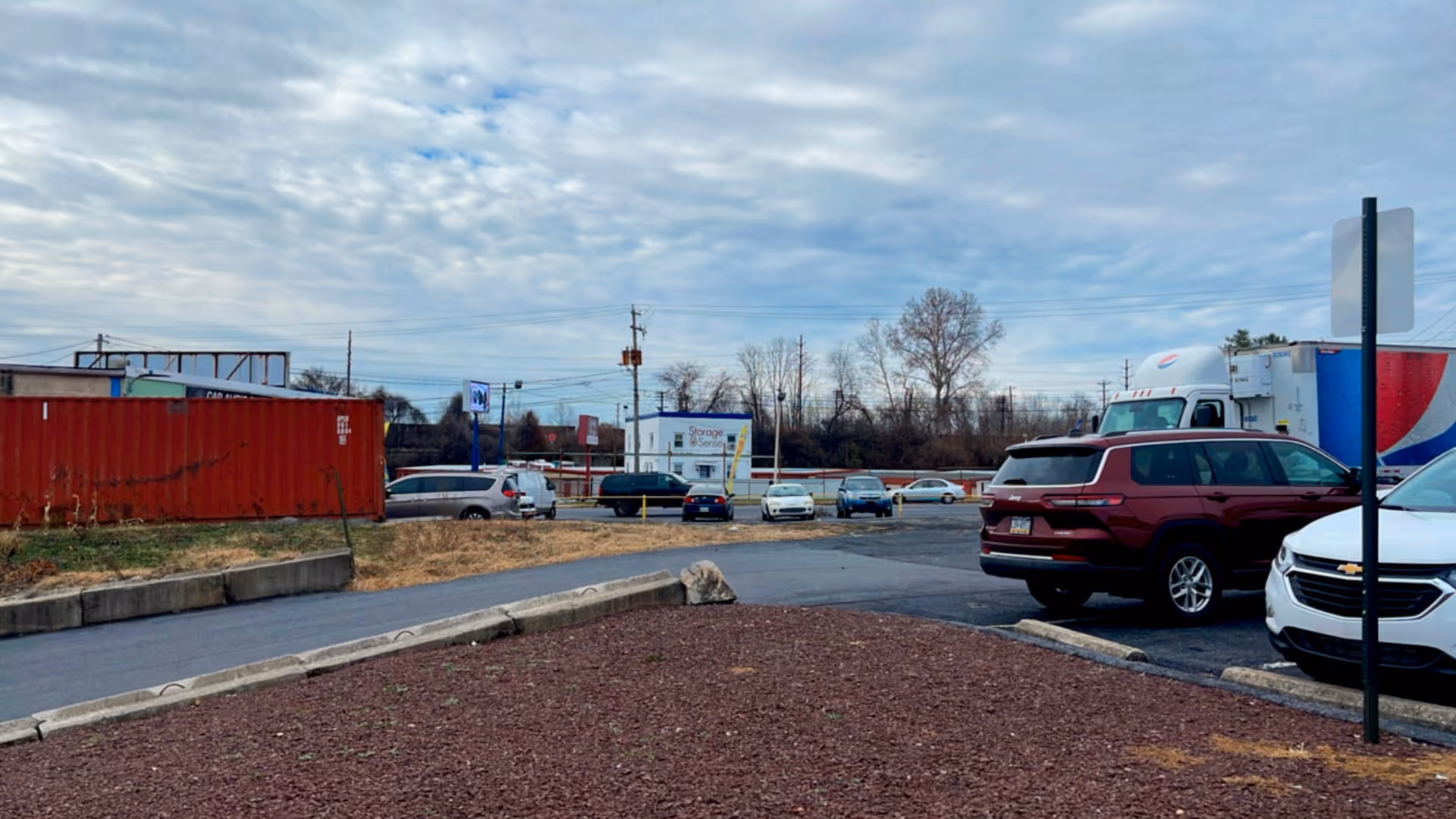 Parking lot view with cars, a red shipping container, and a cloudy sky.