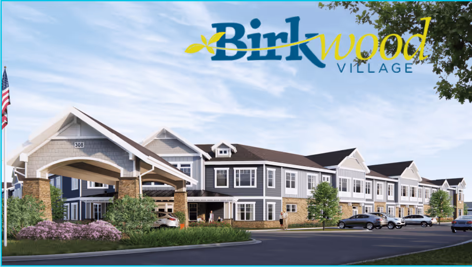Exterior view of Birkwood Village Tama-Toledo, a two-story senior living facility with a covered entrance supported by stone pillars, landscaped greenery, parked cars, and an American flag on the left side under a partly cloudy sky.