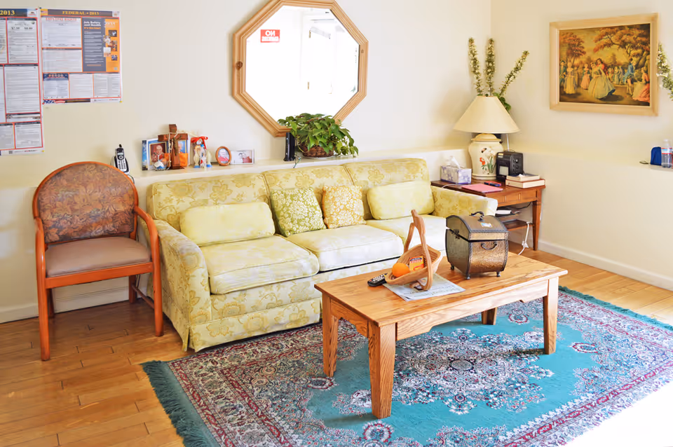 A cozy living room with a floral patterned yellow sofa adorned with several cushions, a wooden coffee table with a basket of fruit and a decorative box, a patterned blue area rug, a wooden chair with floral upholstery, a side table with a lamp, books, and a radio, a wall mirror, framed artwork, and some plants and personal items on a shelf.