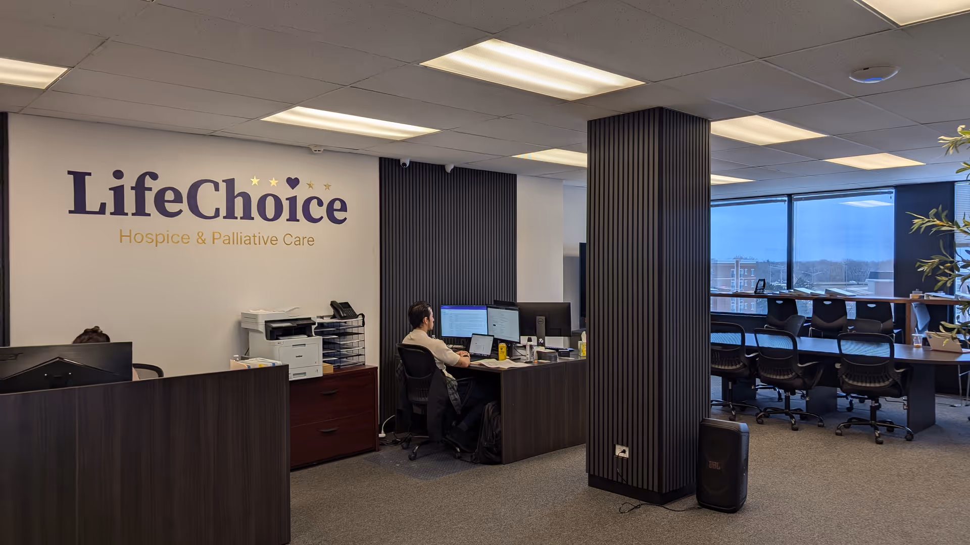Reception area inside LifeChoice Hospice office with desks, computers, and a logo wall.