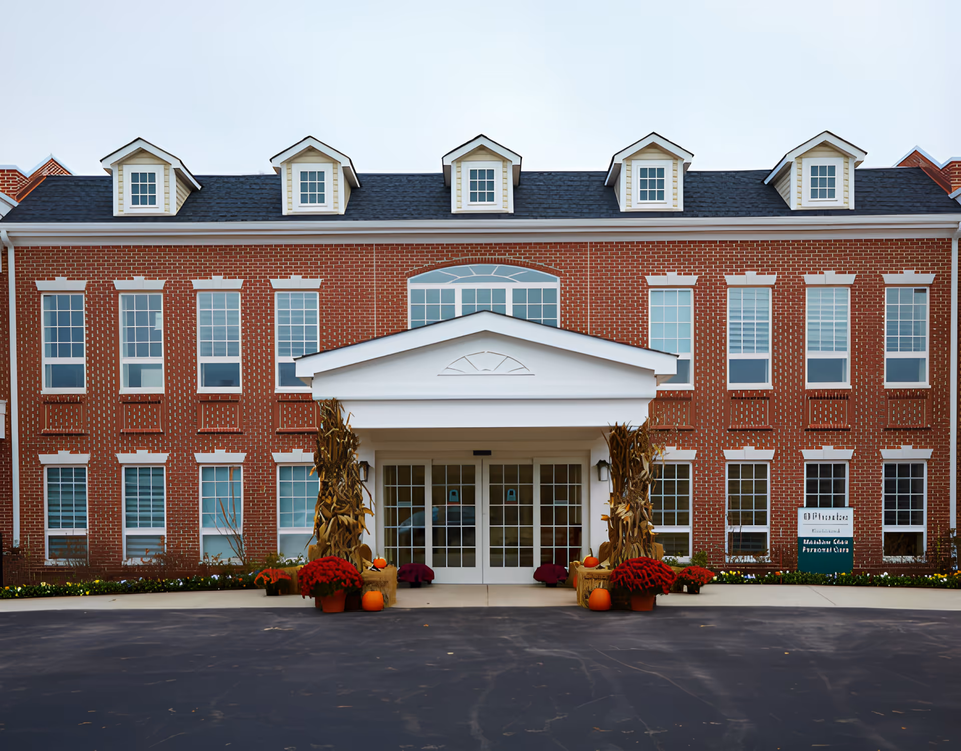 Front entrance of a red-brick building with a white portico, many windows and dormers, and seasonal pumpkins and flowers at the doorway.