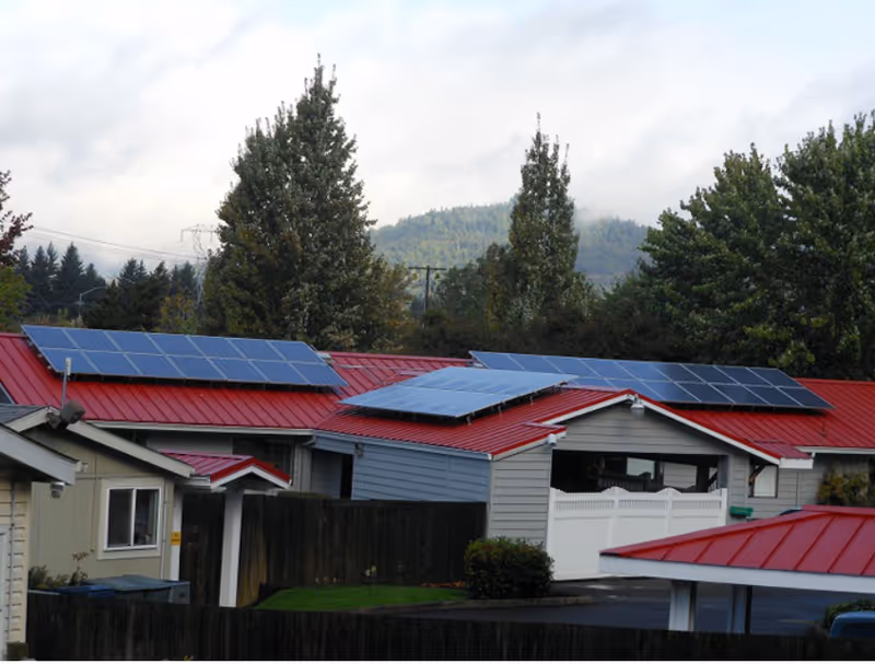 Single-story buildings with red metal roofs fitted with solar panels, a white fence, and trees and hills in the background.