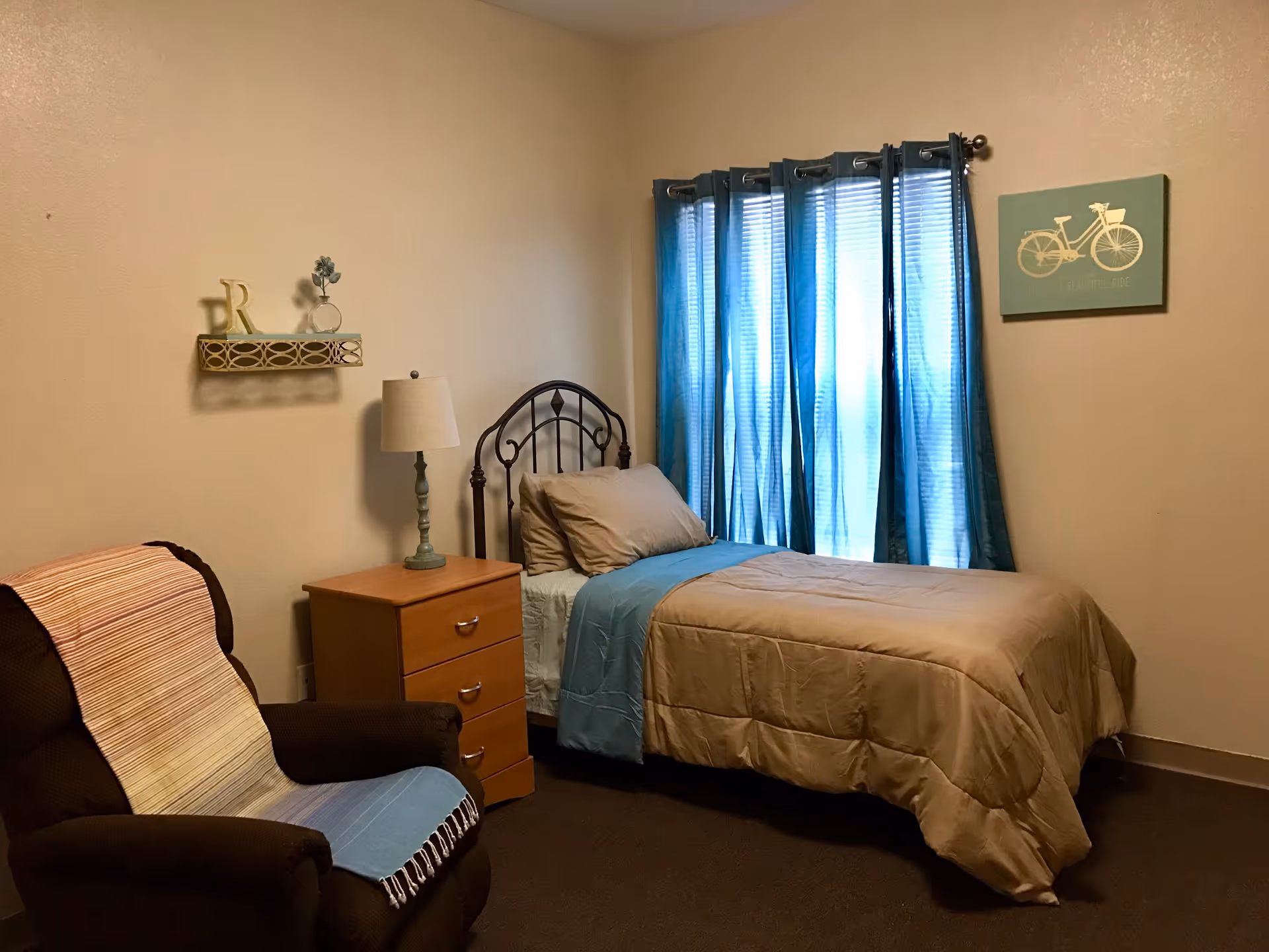 A cozy bedroom with a single bed made with beige and blue bedding, a wooden nightstand with a lamp, a brown armchair with a striped throw blanket, blue curtains covering a window, and a wall decoration featuring a bicycle.