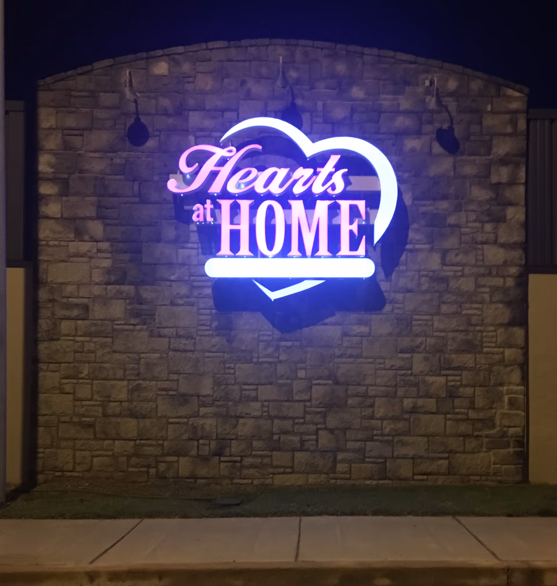 Night exterior of a stone building with a glowing Hearts at Home sign on the front.