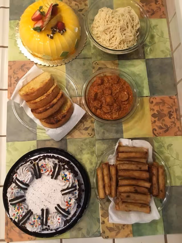 A table with various food items including a yellow fruit-topped cake, a bowl of plain noodles, a bowl of meatballs in sauce, slices of toasted bread on a napkin, a cookies and cream cake with whipped cream and sprinkles, and a plate of fried spring rolls.