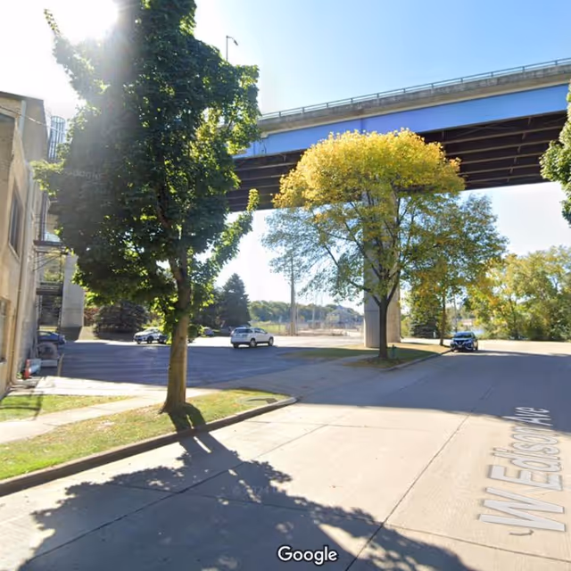 Outdoor street scene with trees, a bridge overhead, and passing cars.