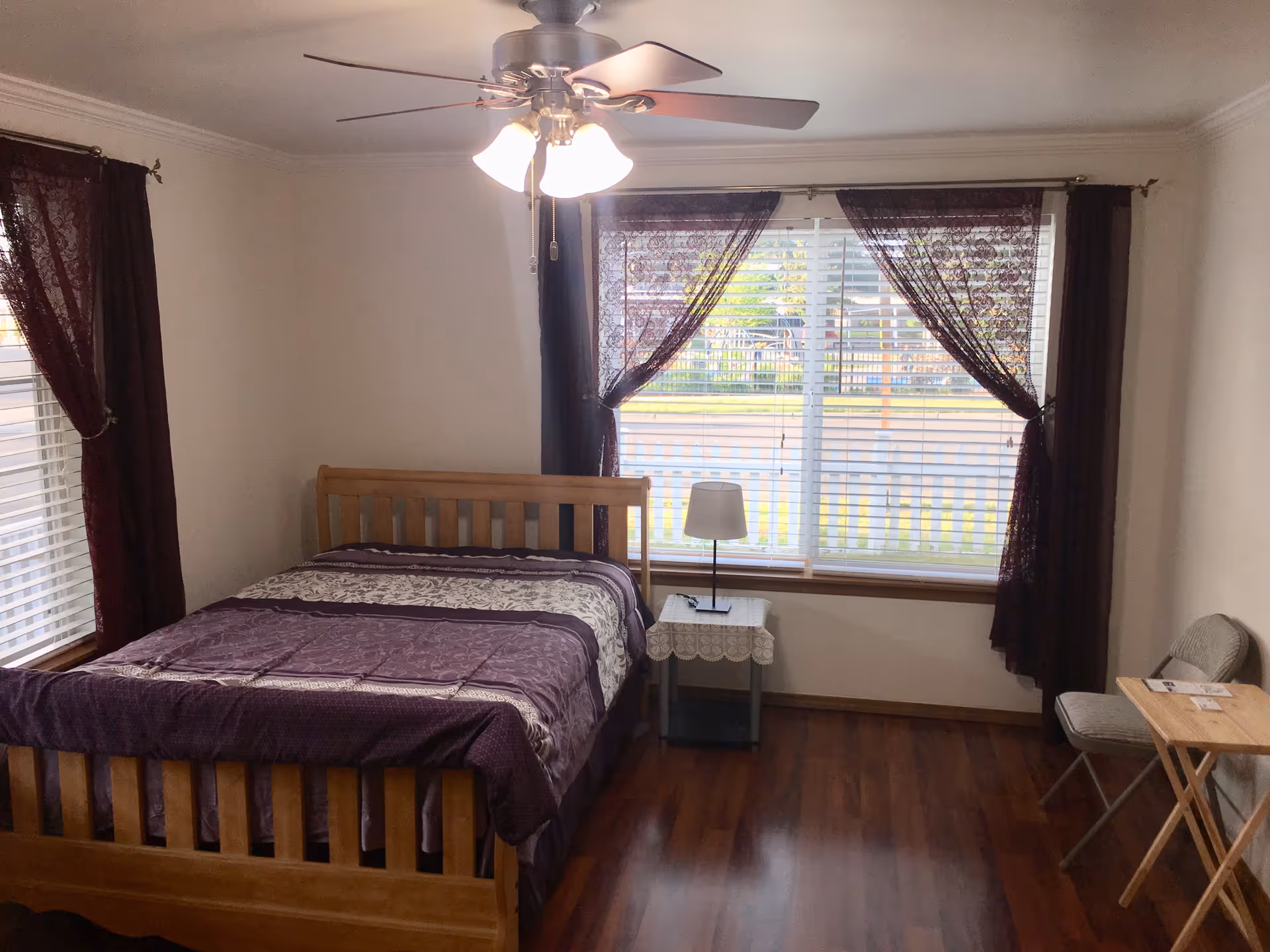 A bedroom with a wooden bed frame and a purple patterned bedspread. There are two windows with white blinds and dark purple curtains. A small table with a lamp is placed between the windows. A folding chair and a small wooden folding table are positioned against the right wall. The room has wooden flooring and a ceiling fan with lights.