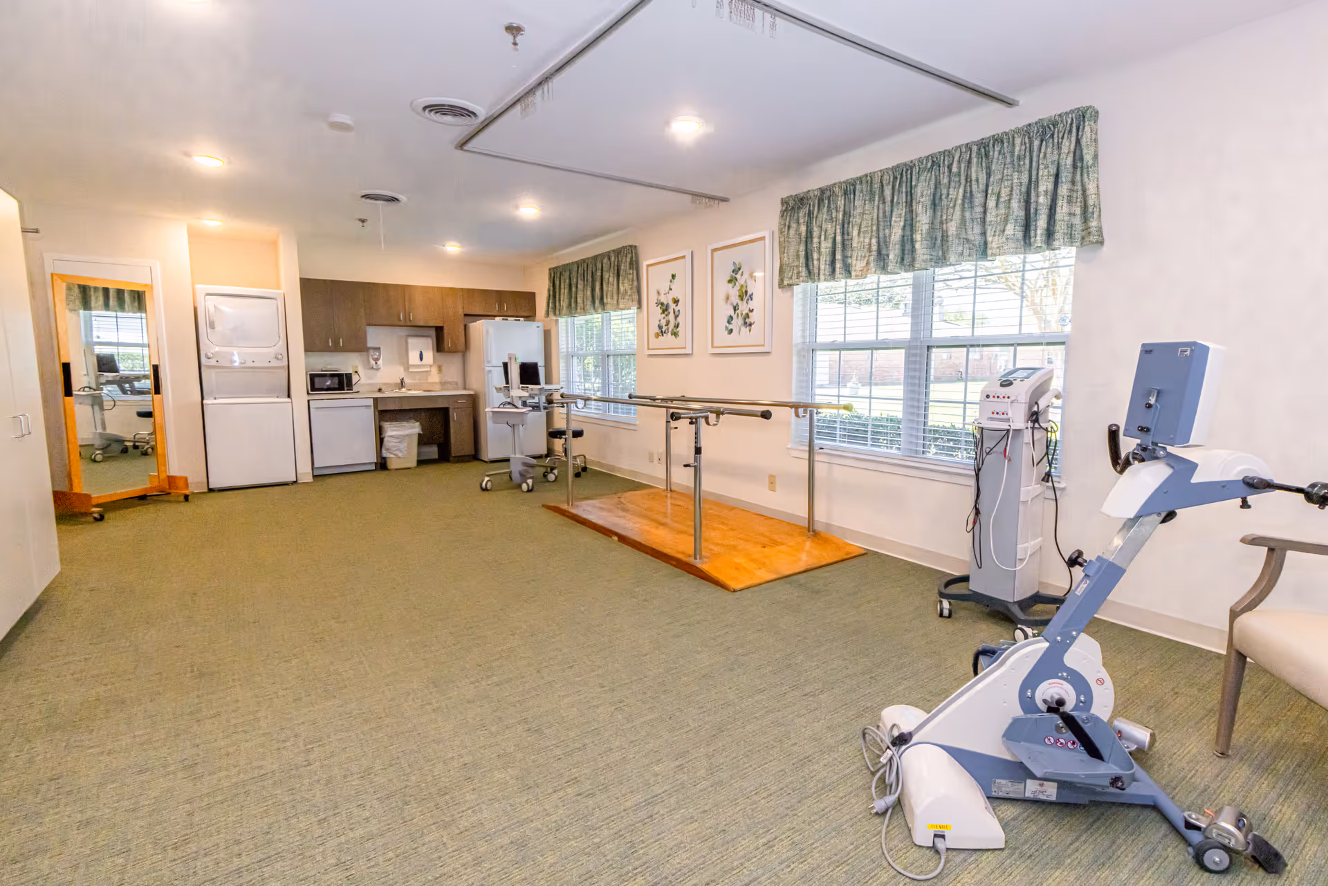 Interior senior living gym with parallel bars, exercise bike, and kitchenette.