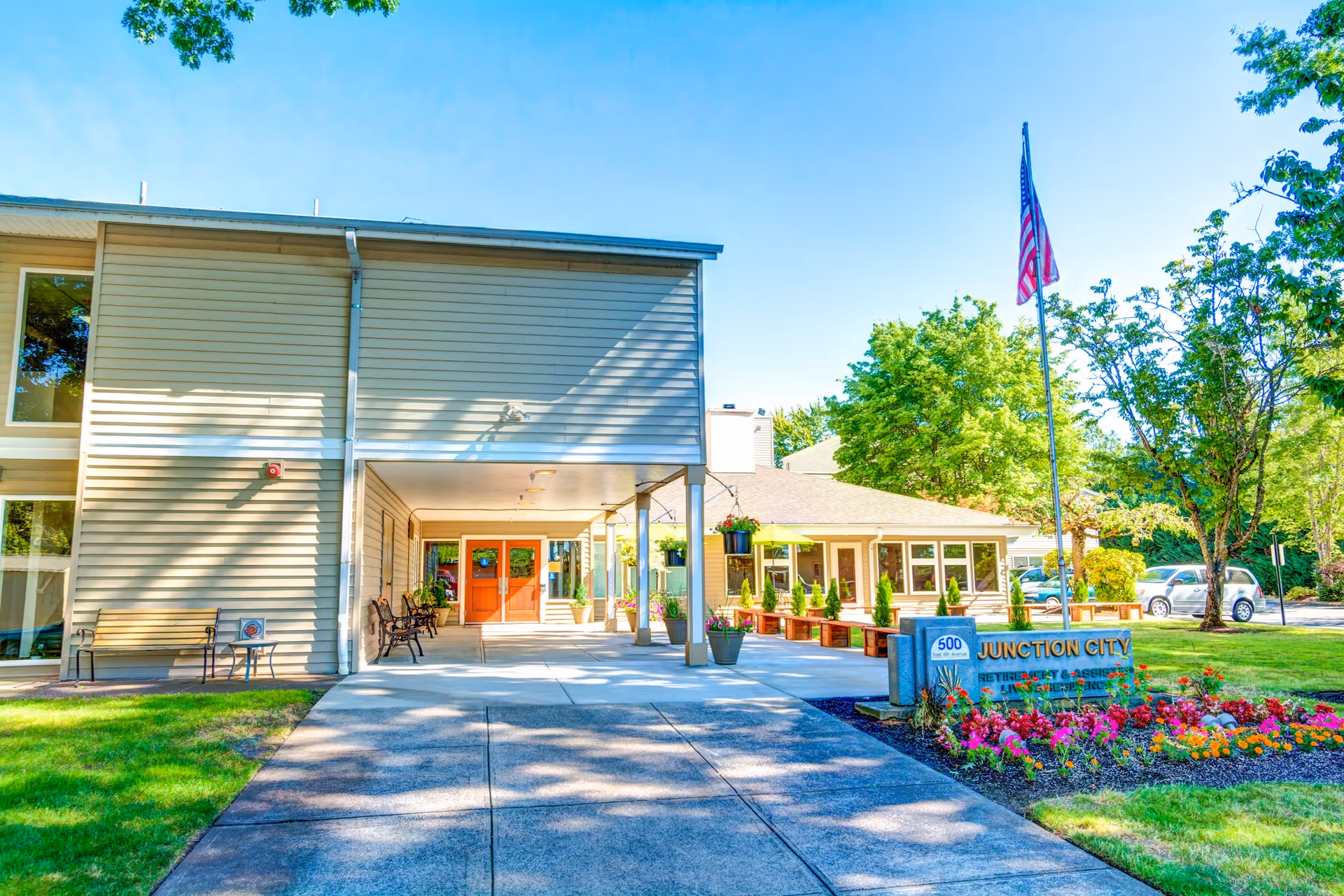 Front entrance of Junction City Retirement and Assisted Living with a covered walkway, flagpole, and flowerbeds.