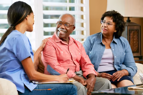 Caregiver chats with smiling elderly couple in a living room.