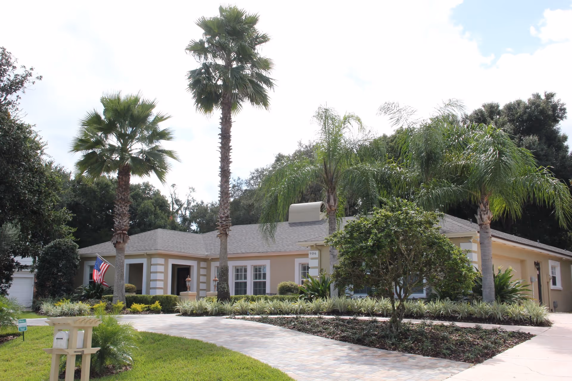 Exterior view of a single-story residential building with a beige facade, surrounded by palm trees and other greenery. A paved driveway leads up to the entrance, and an American flag is displayed near the front door. The sky is partly cloudy.
