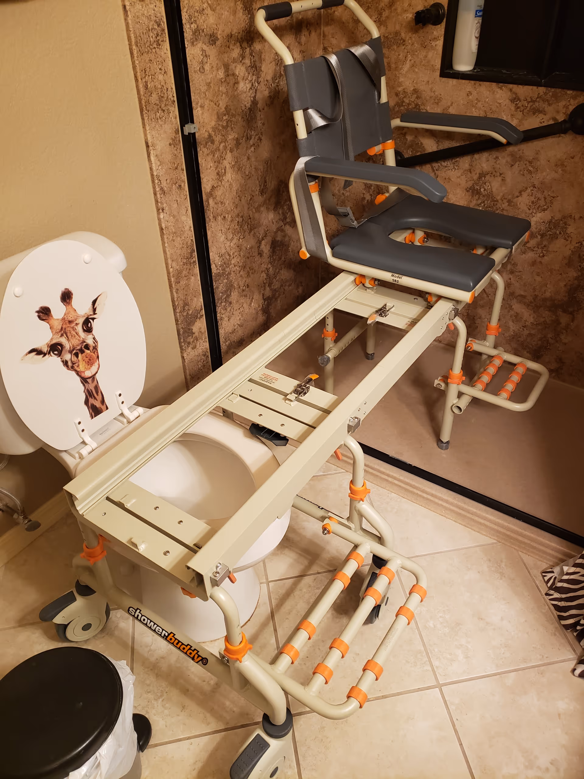 Giraffe-themed toilet seat with a transfer chair over a tiled bathroom.
