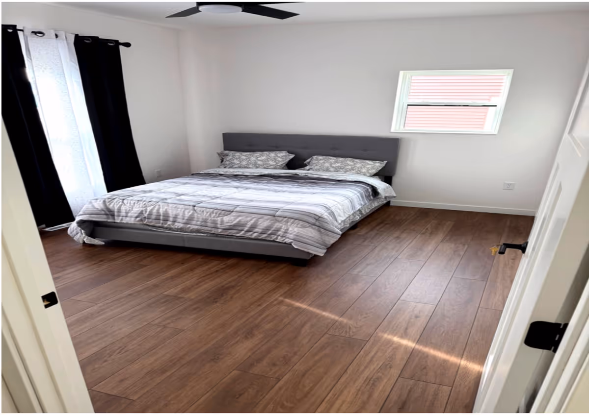 Modern bedroom with gray upholstered bed, wood-look flooring, window, and ceiling fan.