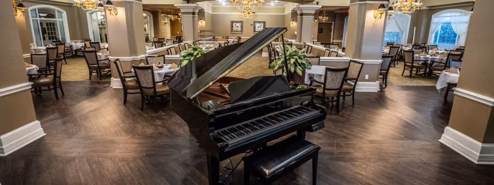 Interior view of a spacious dining room with multiple tables covered in white tablecloths and surrounded by chairs. A black grand piano is prominently placed in the center of the room. The room features wooden flooring, chandeliers hanging from the ceiling, and large windows with curtains allowing natural light to enter.