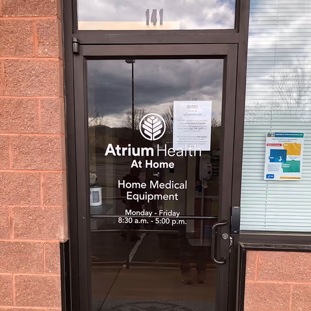 Glass front door shows Atrium Health At Home Home Medical Equipment signage on brick building exterior.