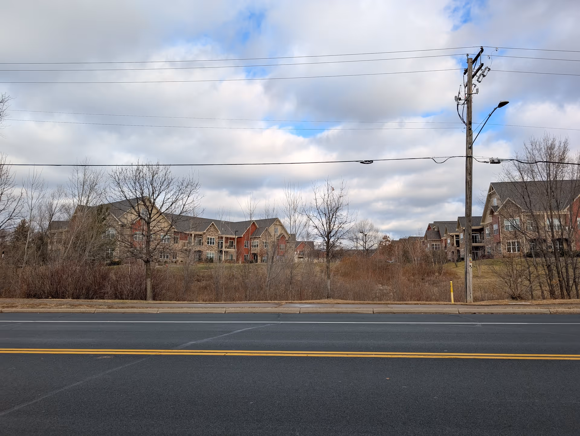 View across a road of a multi-story senior living complex with leafless trees, power lines, and a cloudy sky.