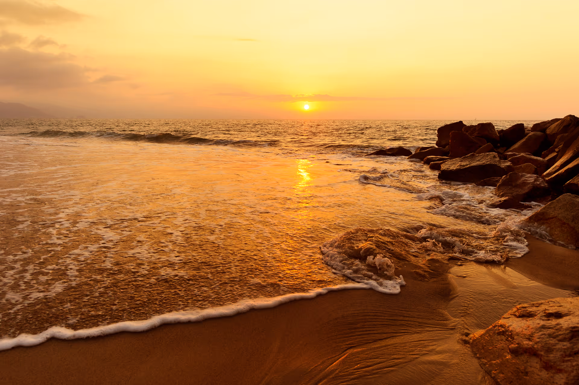 Sunset over the ocean with waves, rocky shoreline, and sandy beach.