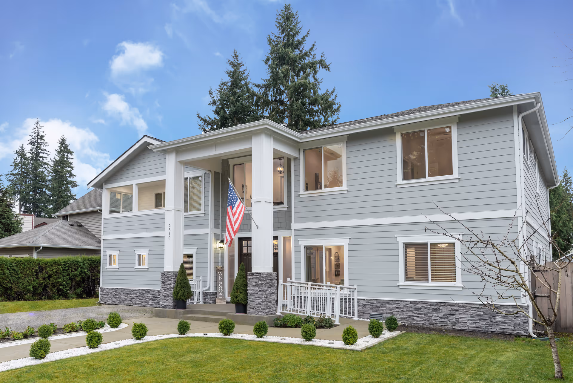 Front exterior view of a two-story senior care facility named Misty Meadows Senior Care, featuring light gray siding with white trim, a covered entrance with columns, an American flag, and a well-maintained lawn with small shrubs and a concrete walkway.