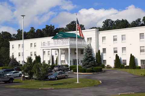 White three-story senior living building with a green awning over the entrance, parked cars, and an American flag in front.