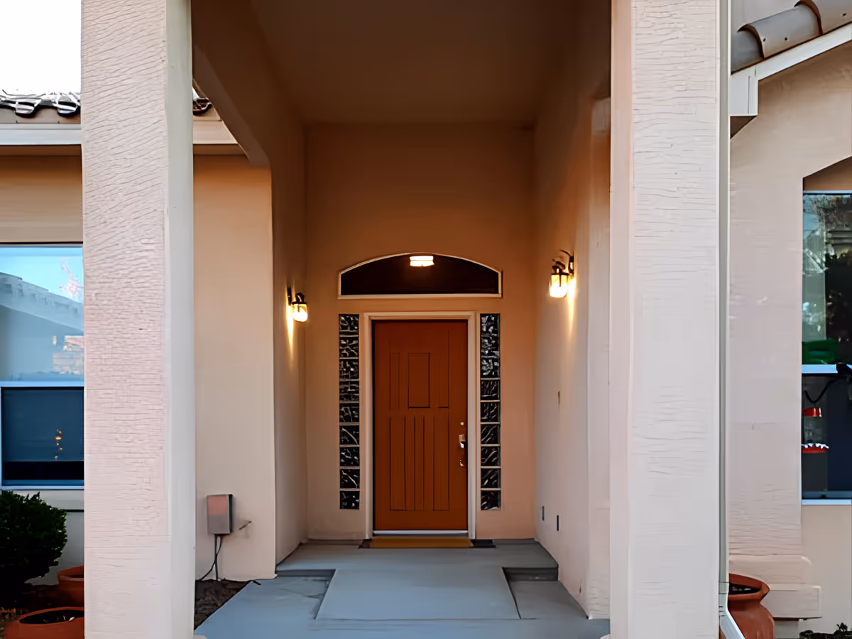 Front entrance of a building with a brown door framed by glass blocks on either side, two wall-mounted lights on each side of the door, and a covered porch area with beige walls and columns.