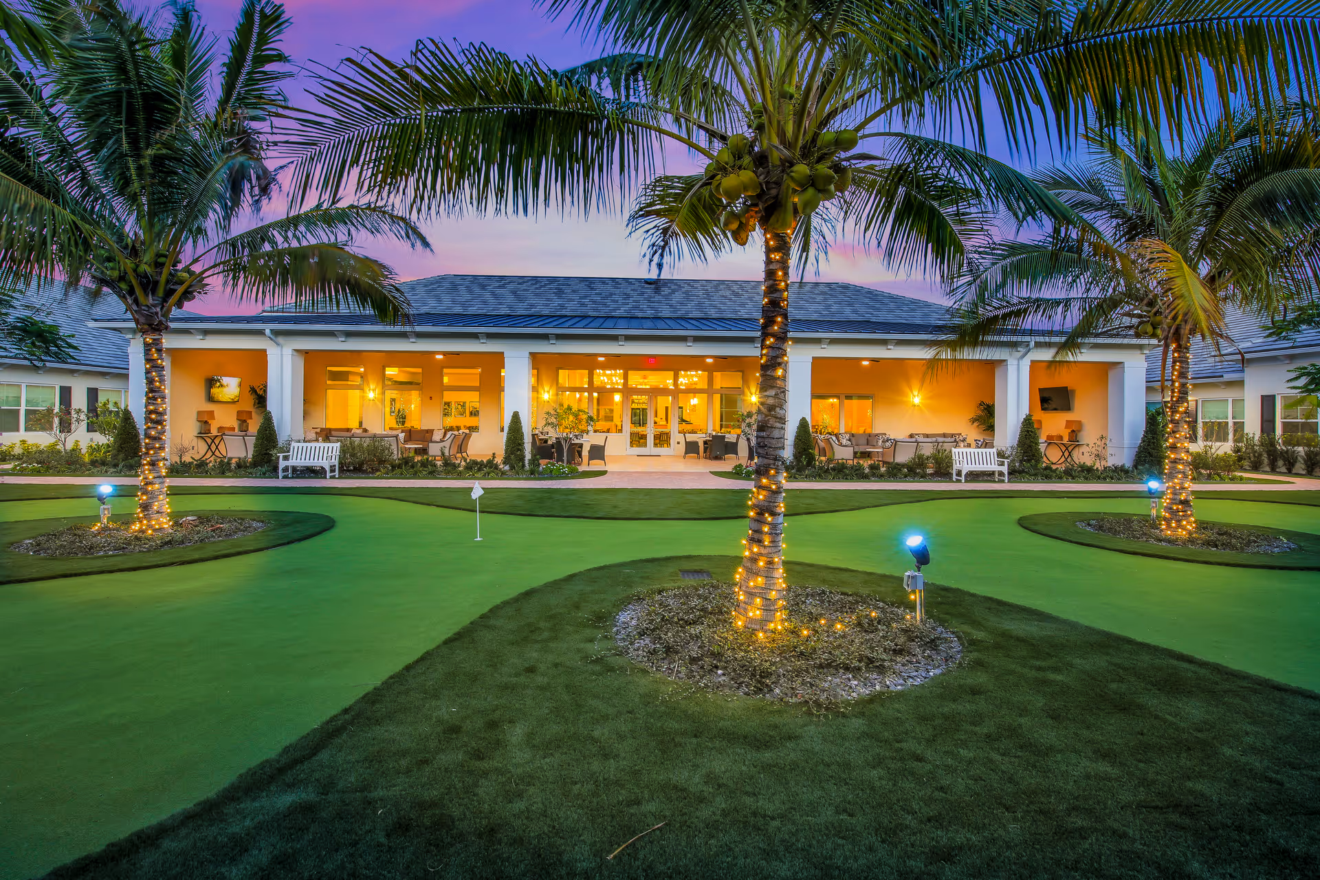 Evening view of a senior living facility with a putting green in the foreground, palm trees wrapped with string lights, and a well-lit building with outdoor seating areas in the background under a purple and blue sky.