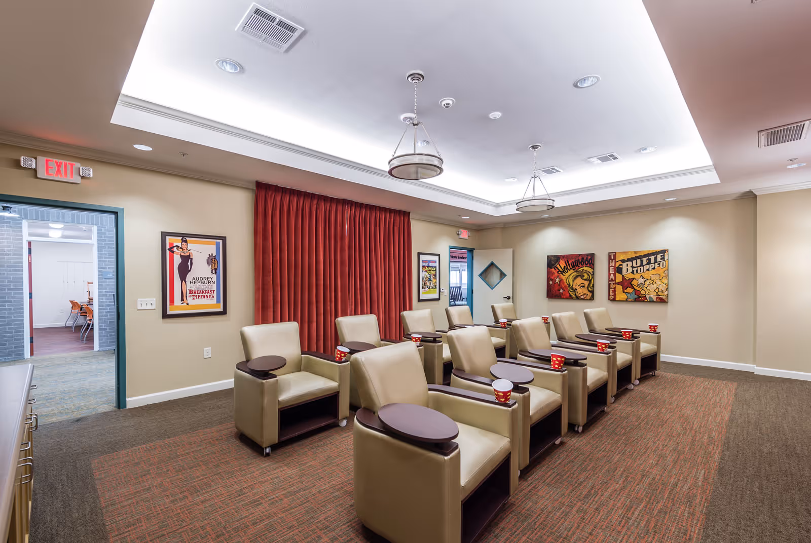 A small theater room with two rows of beige leather recliners, each equipped with a small round table and a red cup. The room has a recessed ceiling with modern light fixtures, red curtains on one wall, and vintage movie posters on the walls. There is an exit door on the left side leading to another room with chairs and tables.