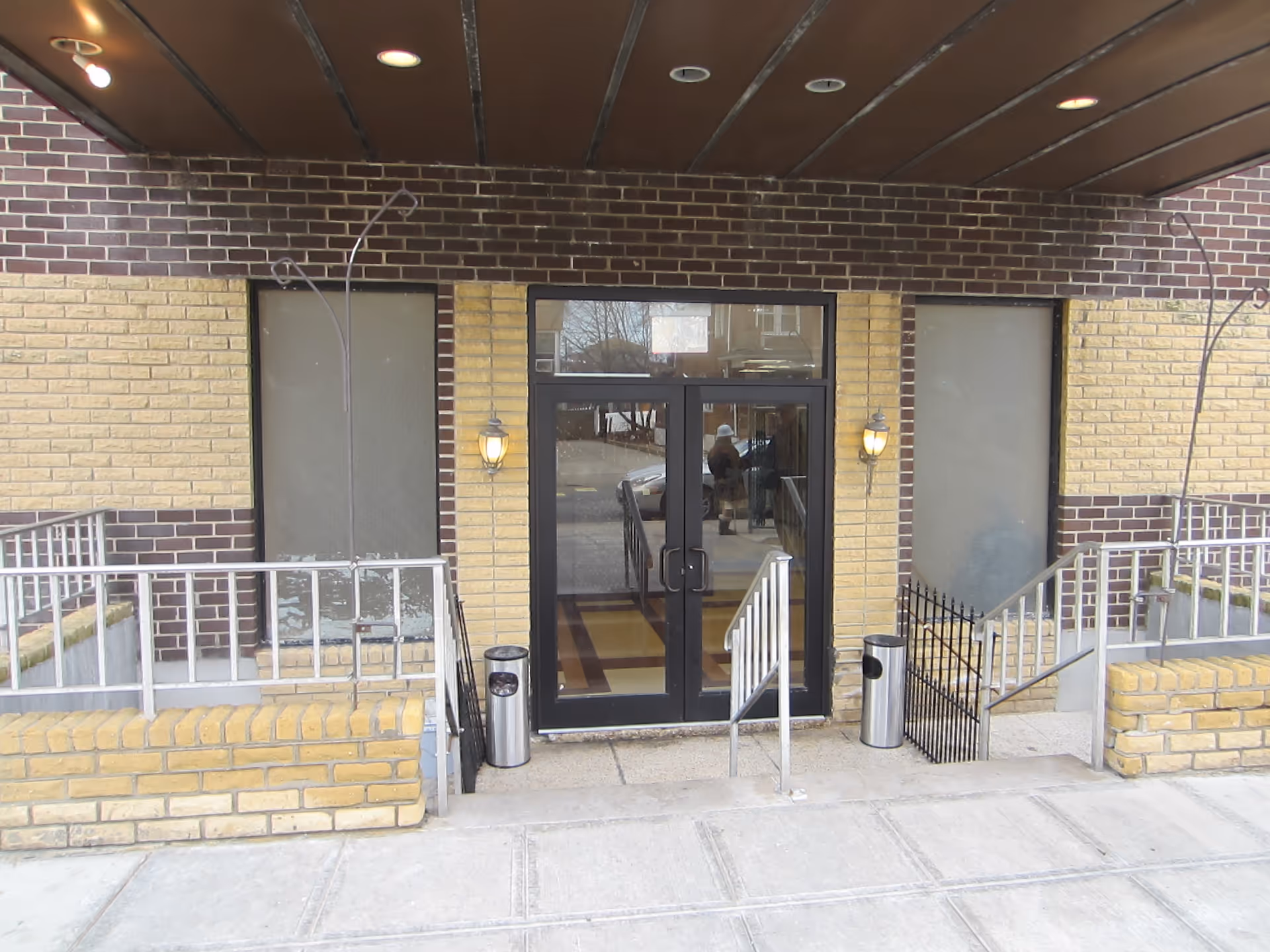 Glass double-door entrance to a brick building with ramps, railings, exterior wall lamps and trash bins.