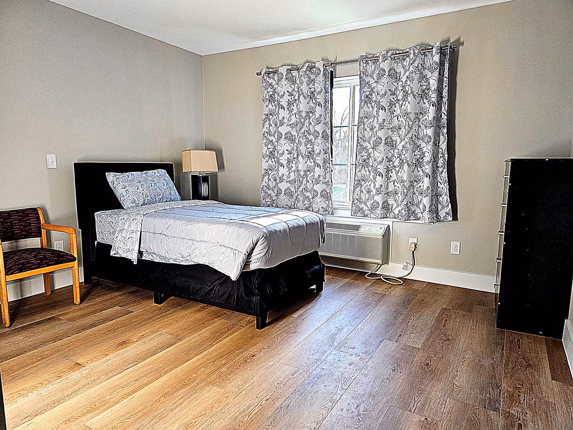 A furnished bedroom features a bed, chair, dresser, and window with floral curtains.