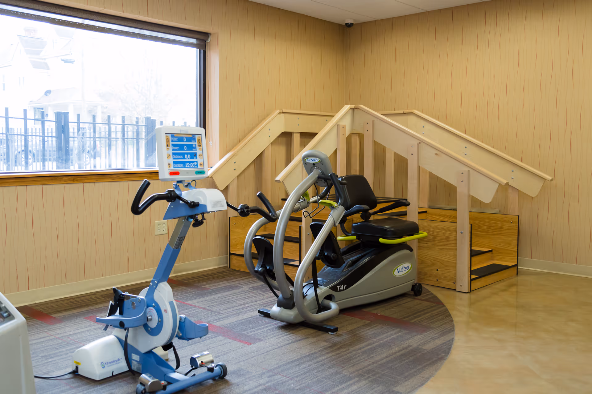 Interior rehabilitation room with two exercise machines and a wooden set of therapy stairs by a window.