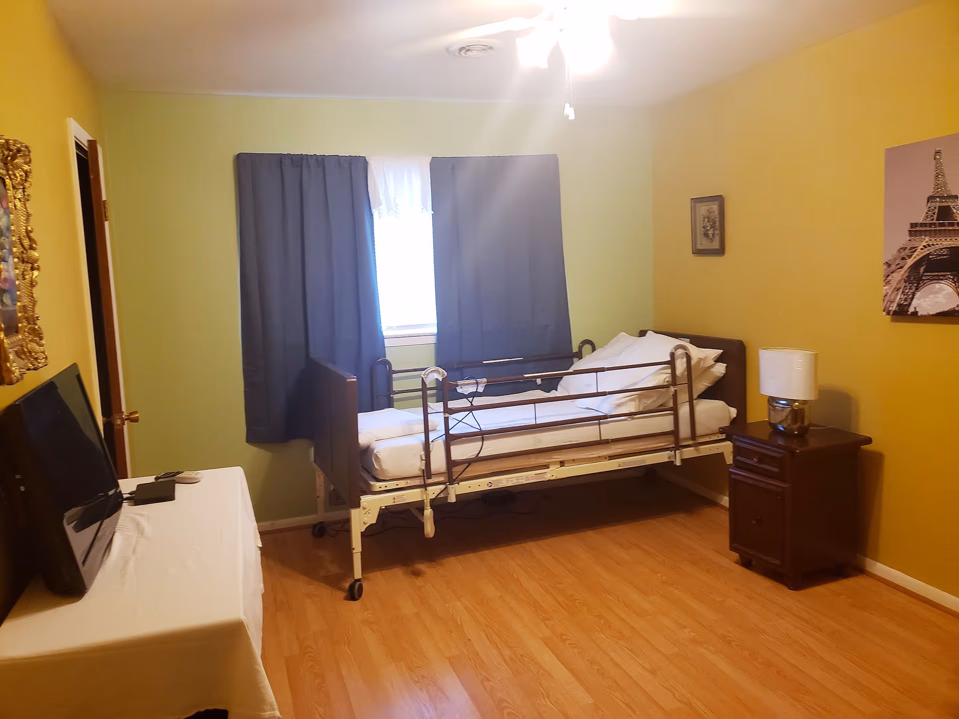Yellow-walled bedroom with metal hospital bed, blue curtains, and a nightstand.
