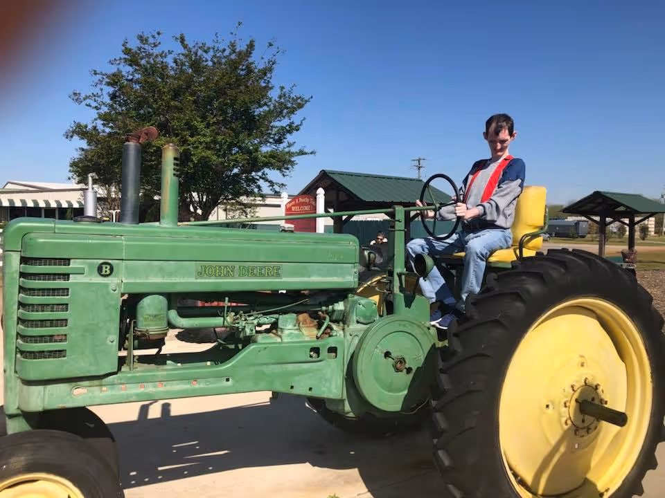 A man sits on a vintage green John Deere tractor outdoors at a facility.