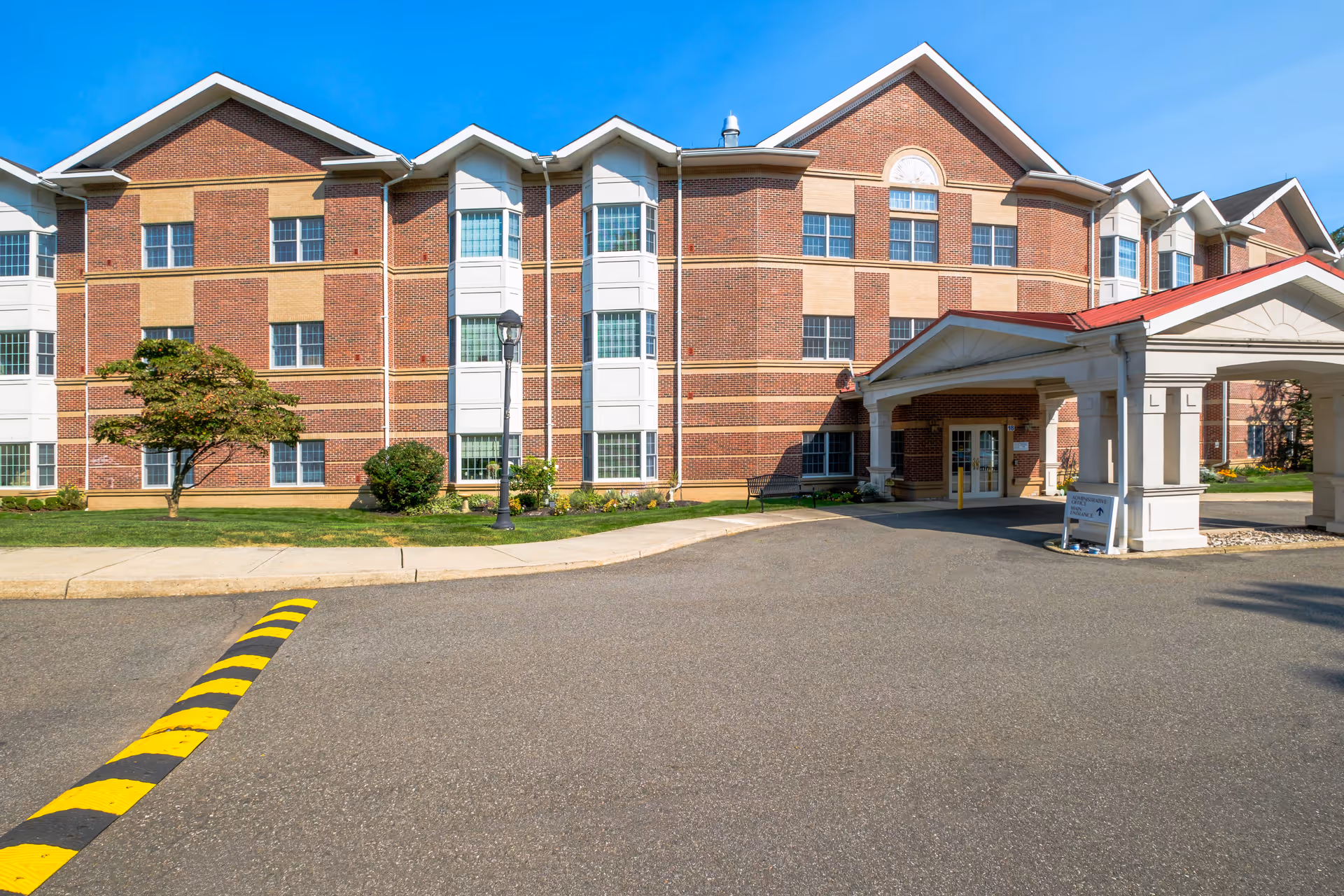 Front entrance of a multi-story brick senior living facility with a covered porte-cochère and driveway.