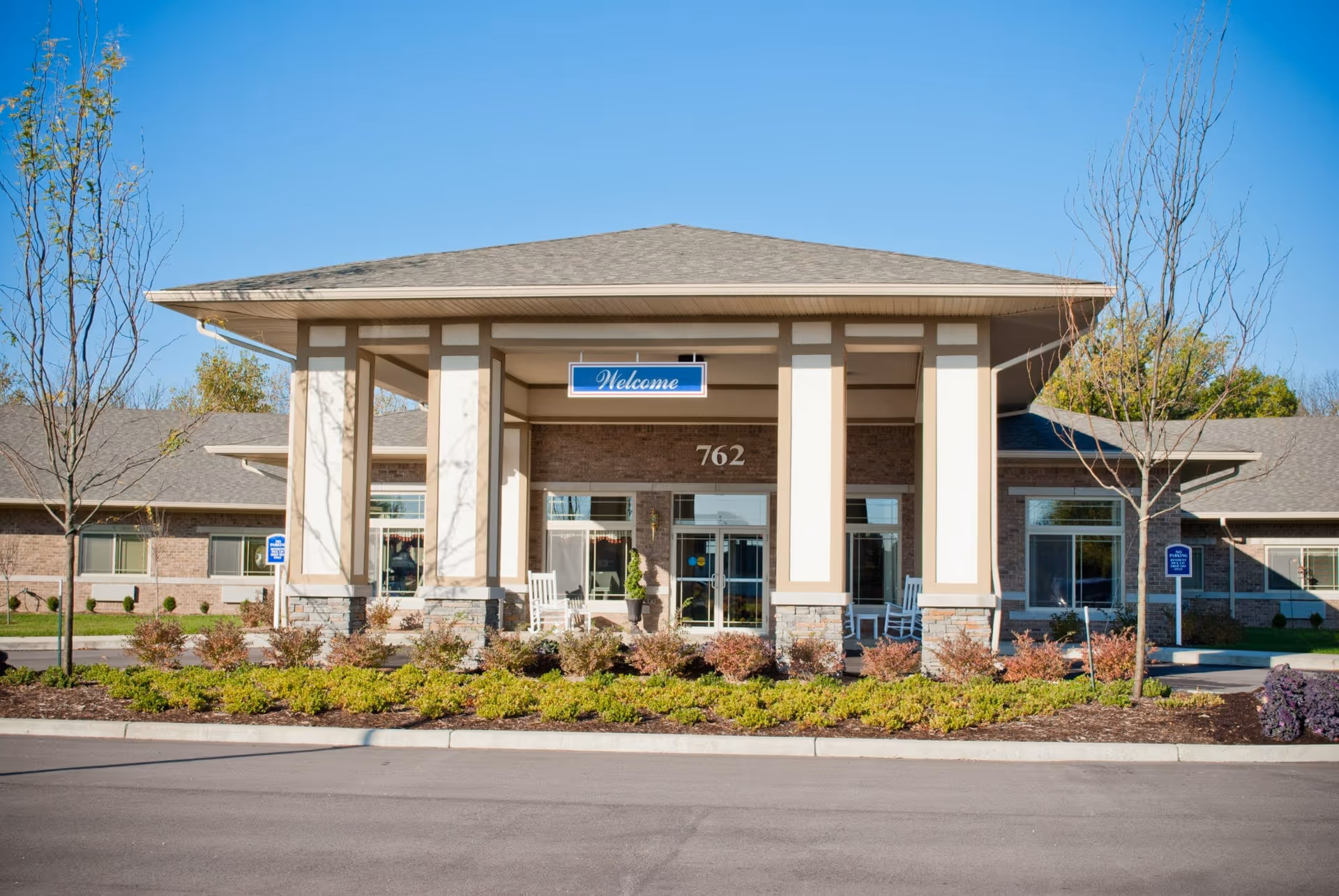 Front entrance of a single-story senior living facility with a covered portico, columns, a 'Welcome' sign and the address number 762, surrounded by landscaping.