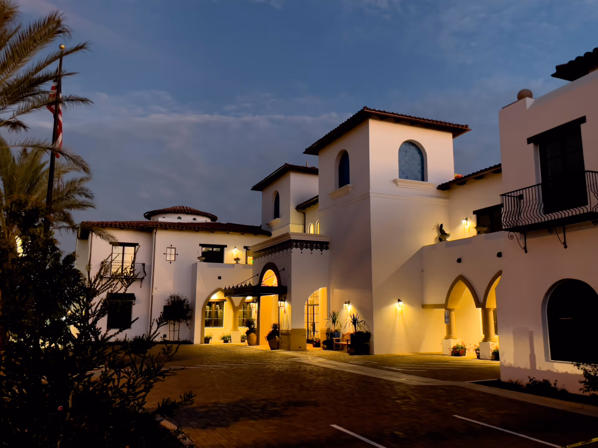 Exterior view of a Mediterranean-style building at dusk with warm lights illuminating the entrance and arches, palm trees on the left, and a partly cloudy sky.