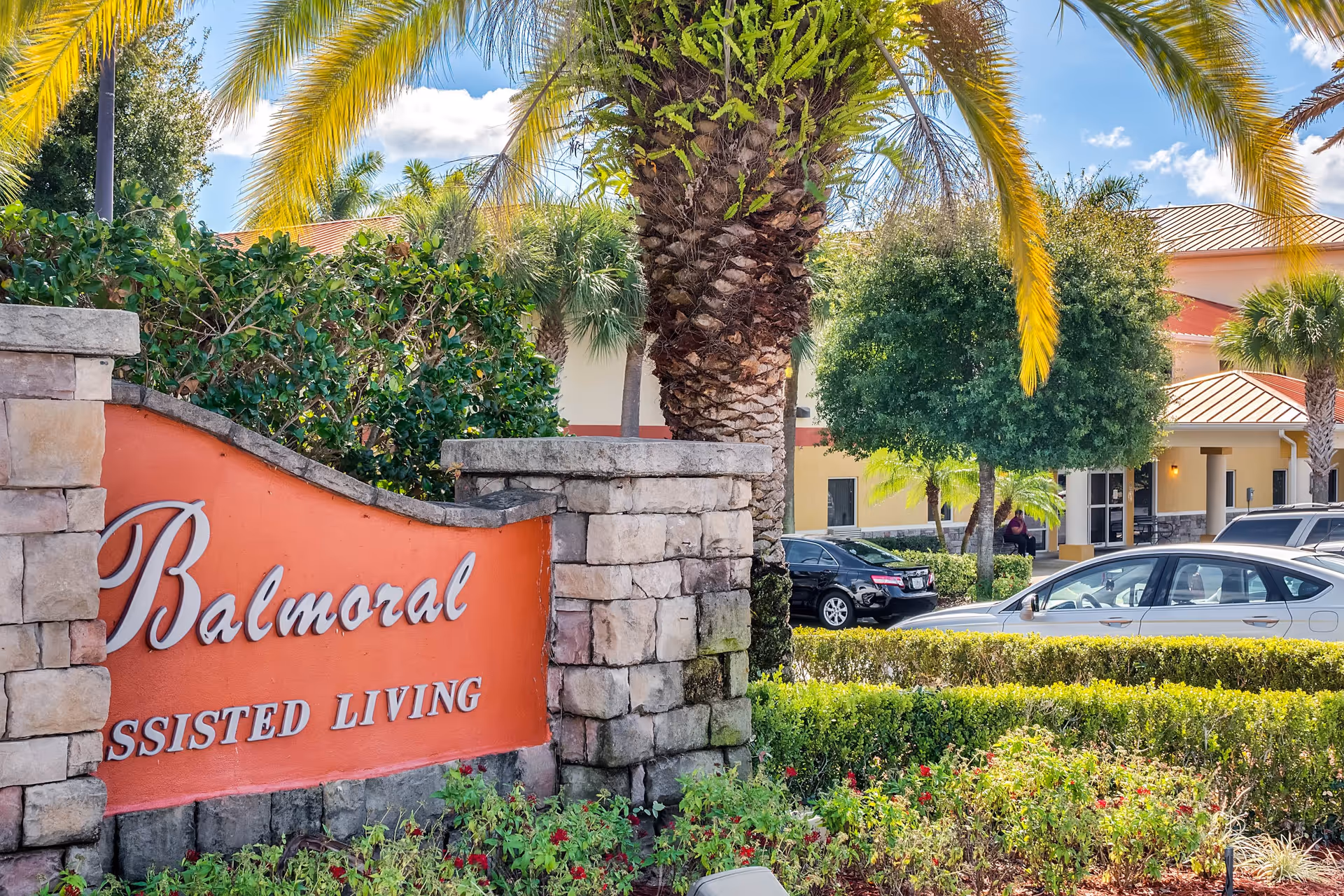 Entrance sign for Balmoral Assisted Living surrounded by greenery and palm trees, with the facility building and parked cars visible in the background under a blue sky with some clouds.