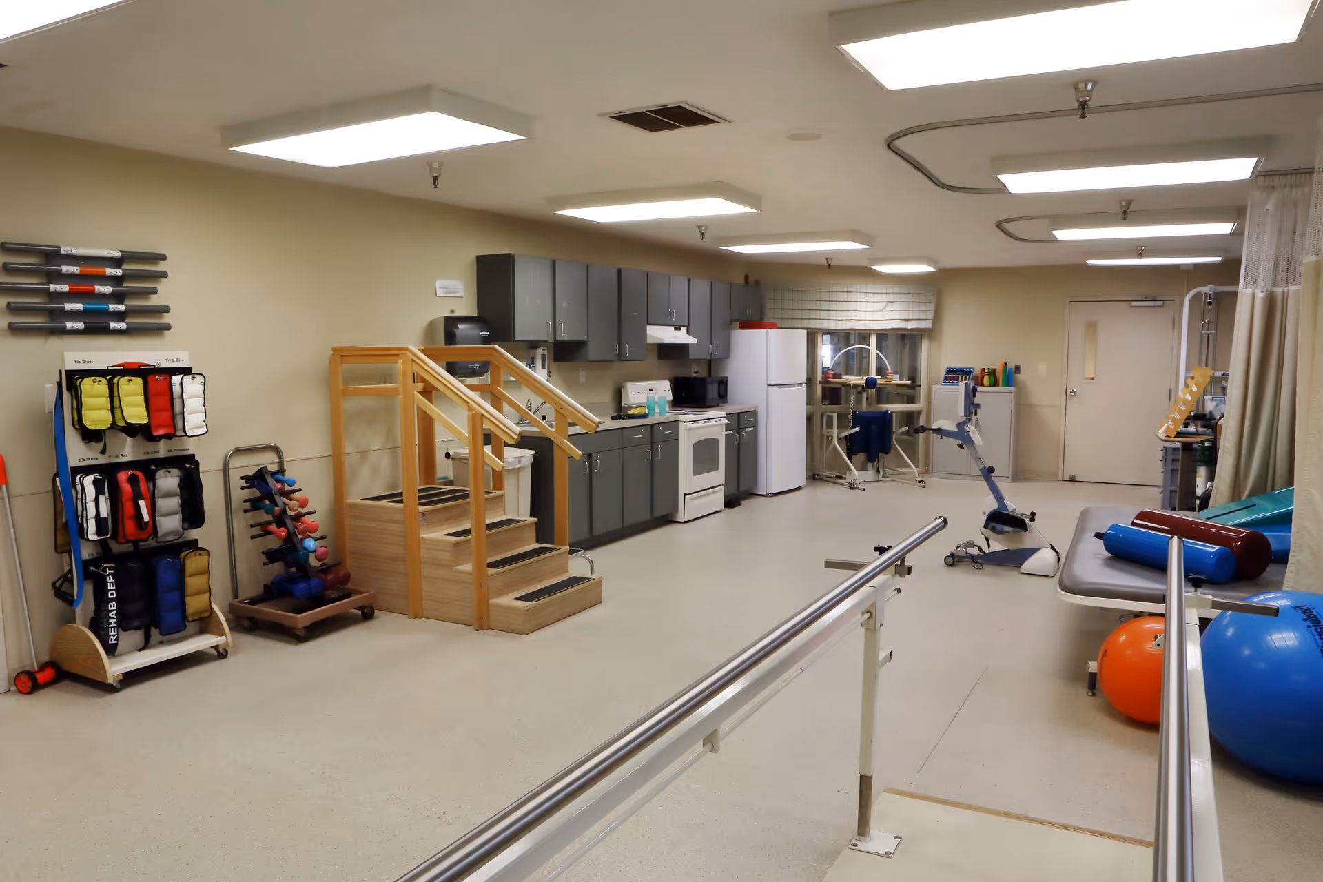 A spacious rehabilitation room with exercise equipment including parallel bars, a small wooden staircase for physical therapy, various weights, exercise balls, and a stationary exercise bike. The room also features a kitchenette area with cabinets, a stove, microwave, and refrigerator. The walls are beige, and the ceiling has multiple fluorescent lights.