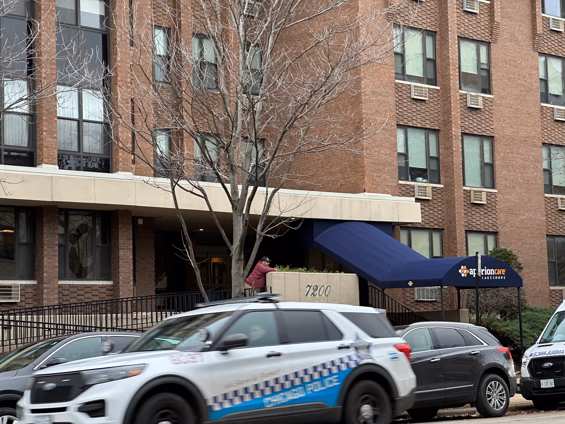 Exterior front entrance of Aperion Care Lakeshore, a brick building with a blue awning, the address '7200', and several parked cars including a police SUV.
