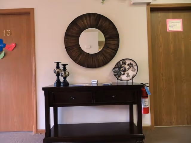 A hallway area with a dark wooden console table against a light-colored wall. On the table are two decorative candle holders and a decorative plate on a stand. Above the table hangs a round mirror with a dark wooden frame. To the left is a wooden door labeled with the number 13 and colorful paper hearts attached. To the right is another wooden door with a small pink sign and a red fire extinguisher mounted on the wall beside it.