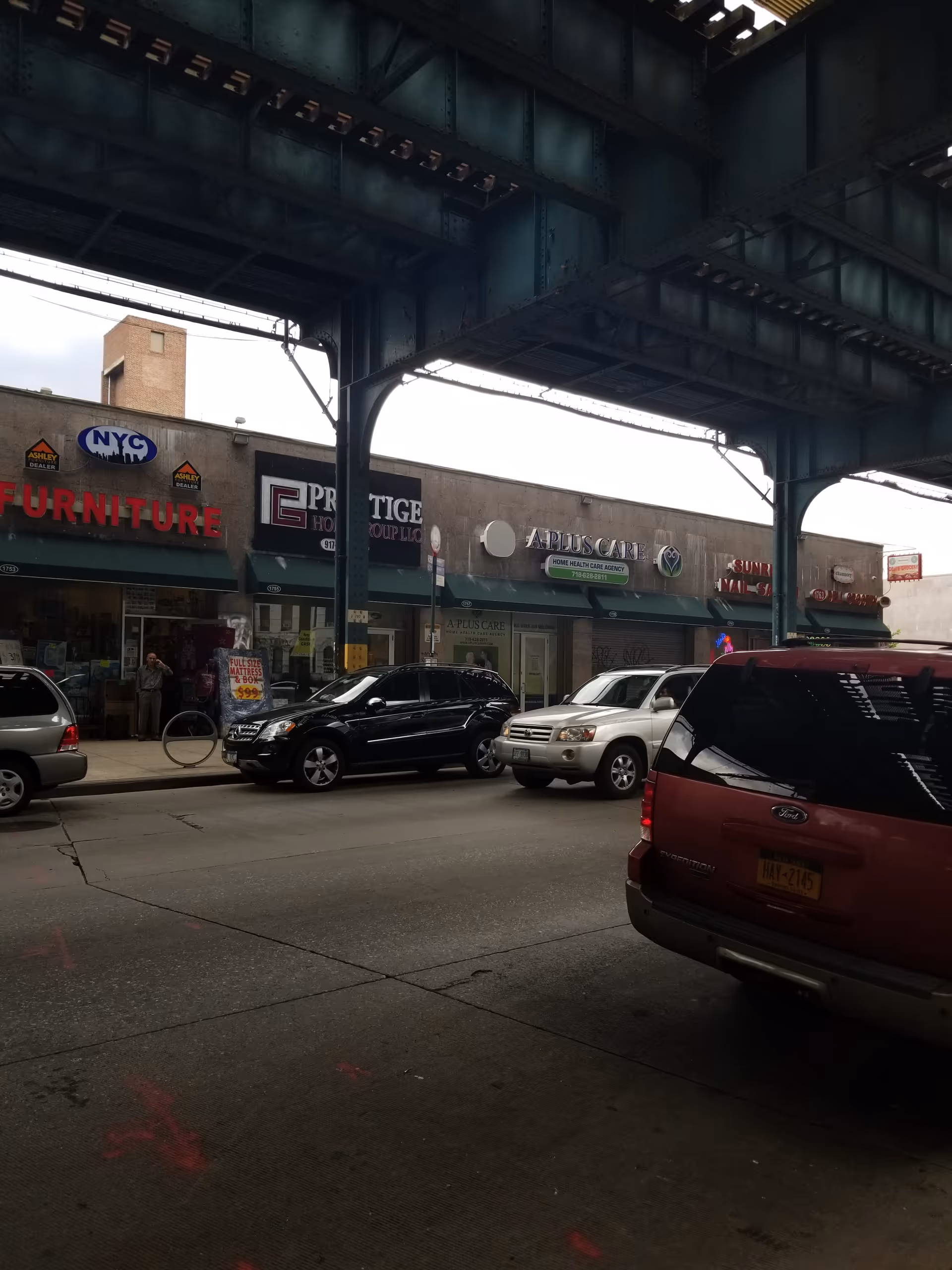 Street-level storefronts under an elevated rail with parked cars along the curb