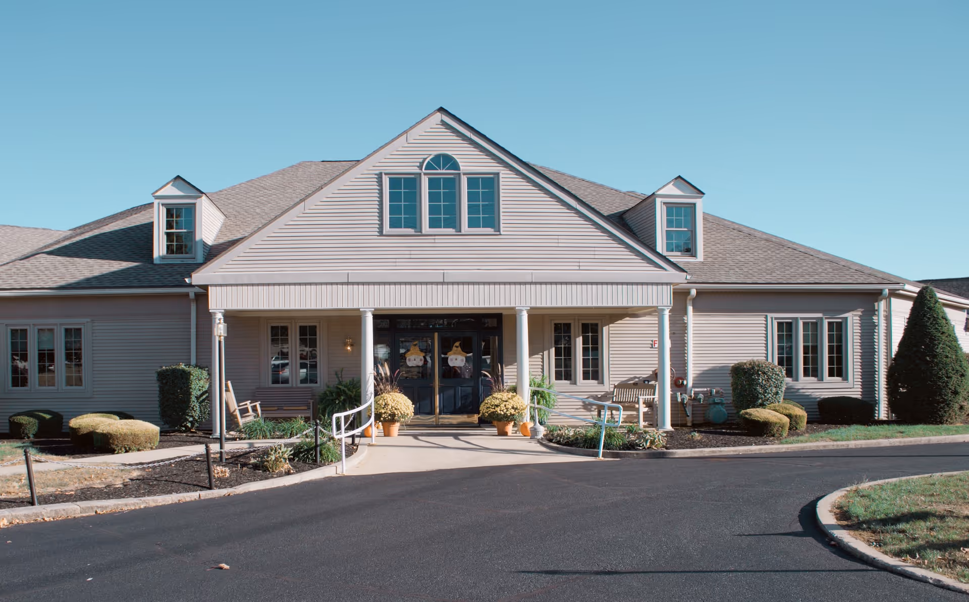 Front exterior view of Windsorwood Place - An Abbington Assisted Living Community building with a paved driveway, landscaped bushes, and a clear blue sky.