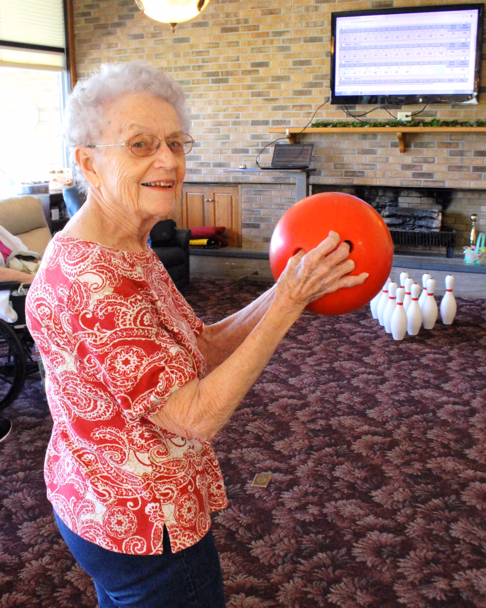 An elderly woman wearing glasses and a red patterned shirt is holding a red bowling ball indoors, preparing to bowl at a set of white pins arranged on a carpeted floor. The room has a brick wall with a mounted TV screen and a fireplace in the background.