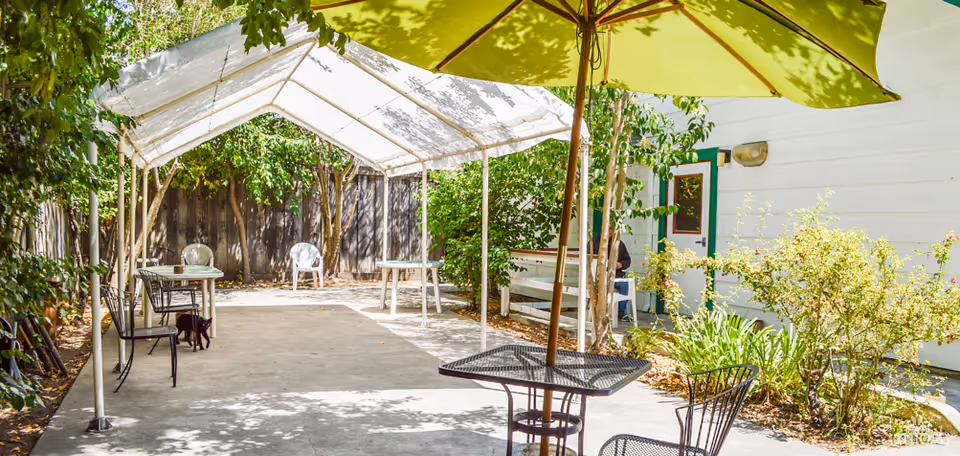 Outdoor patio area with a white canopy tent providing shade over a concrete floor. Several metal chairs and tables are arranged under and around the canopy. A large green umbrella is partially visible in the foreground. The area is surrounded by greenery, including bushes and trees, with a white building wall and door in the background.