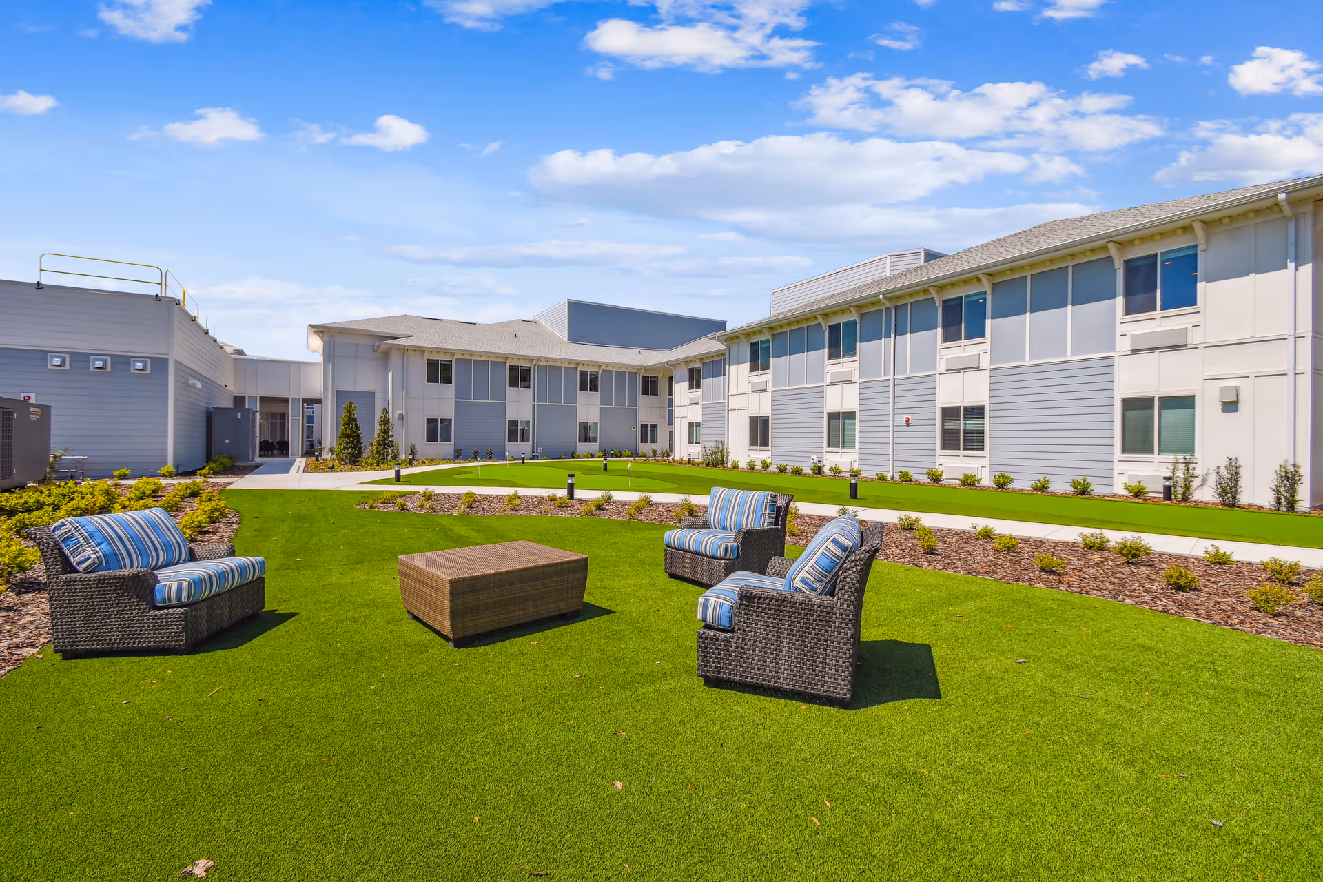 Outdoor courtyard area at HearthStone at Leesburg with green artificial grass, three wicker chairs with blue striped cushions, a wicker coffee table, and a two-story building in the background under a partly cloudy sky.