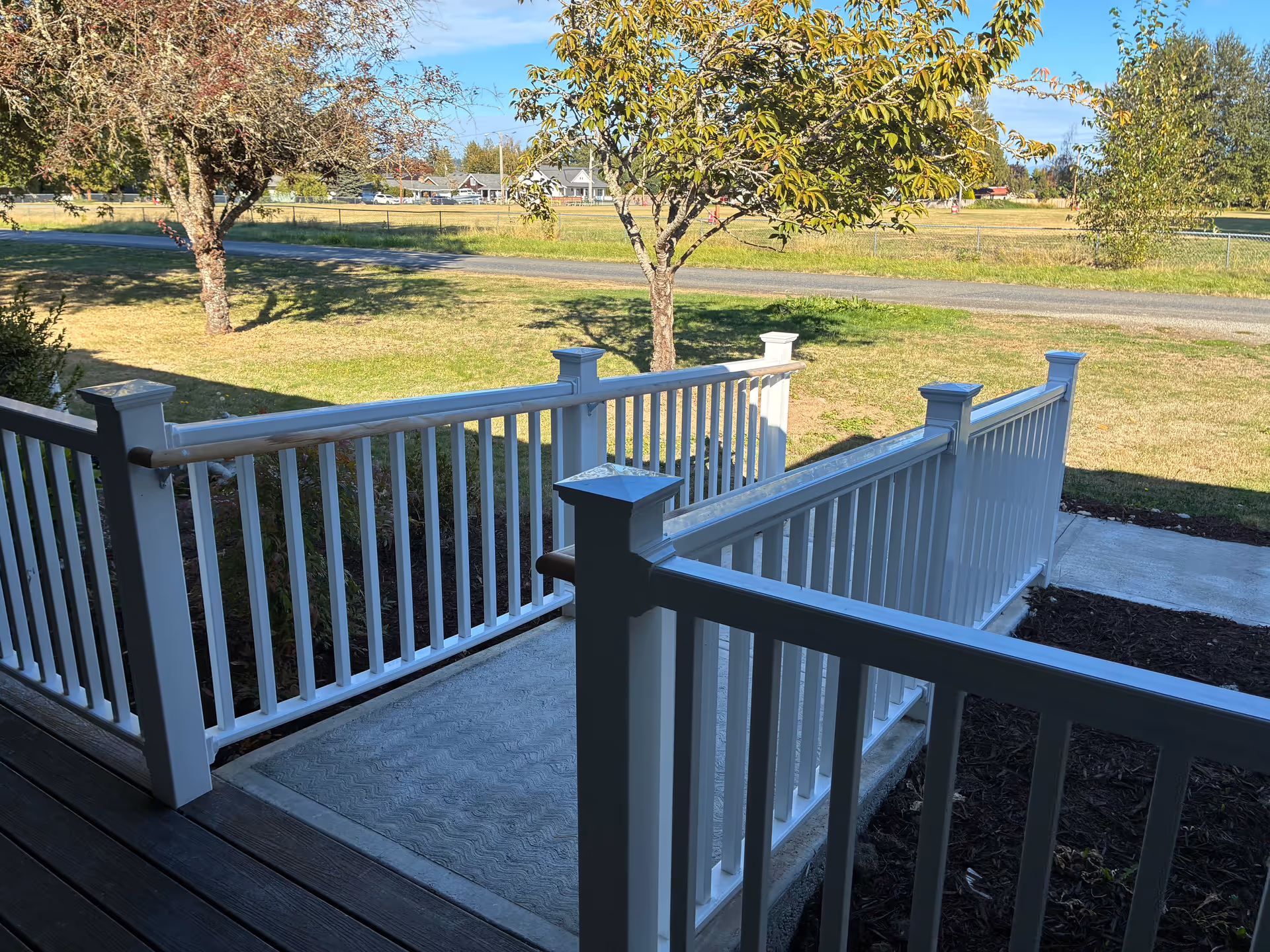 White railing porch overlooking a grassy yard with trees and road.