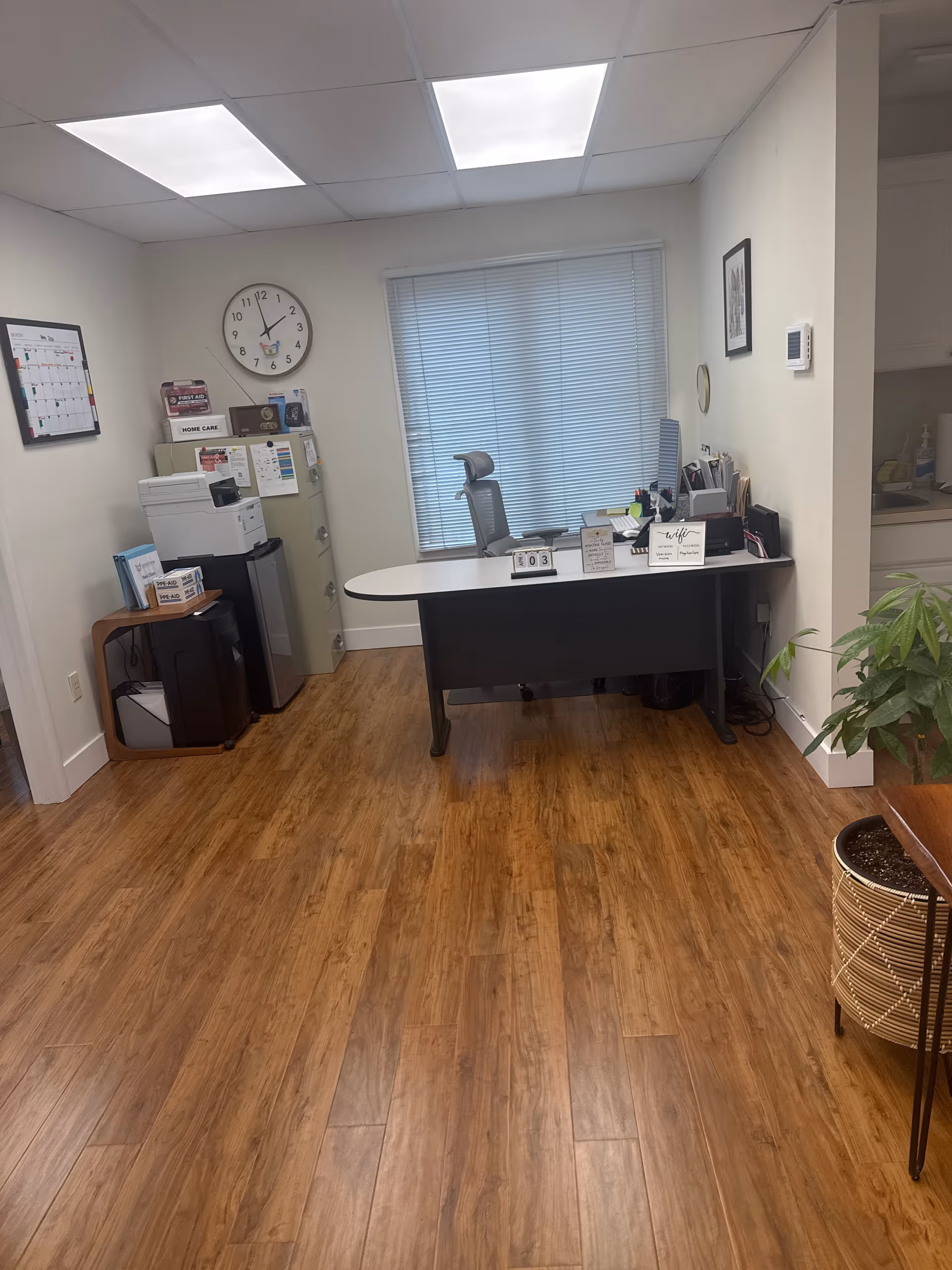 Office desk area in a facility reception with chair, copier, clock, and blinds.